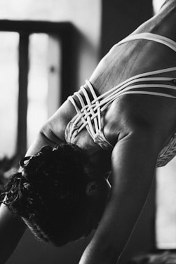 A person is engaged in a yoga or stretching pose, focusing on their back muscles. The intricate design of the straps on their clothing contrasts with the smooth skin. The image is in black and white, providing a dramatic and intimate atmosphere.