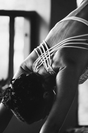 A person is engaged in a yoga or stretching pose, focusing on their back muscles. The intricate design of the straps on their clothing contrasts with the smooth skin. The image is in black and white, providing a dramatic and intimate atmosphere.