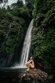 A person in swimwear sits on a rocky outcrop in front of a tall, cascading waterfall surrounded by lush green foliage. The serene setting is enveloped by dense forest, creating a tranquil and natural atmosphere.