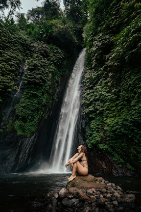A person in swimwear sits on a rocky outcrop in front of a tall, cascading waterfall surrounded by lush green foliage. The serene setting is enveloped by dense forest, creating a tranquil and natural atmosphere.