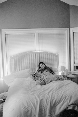 A person is resting on a large, comfortable bed, wrapped in a blanket. The room appears cozy, with closed blinds on the windows behind the bed and soft lighting creating a warm atmosphere. A bedside table with a lamp and some items sits to the side.