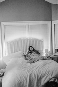 A person is resting on a large, comfortable bed, wrapped in a blanket. The room appears cozy, with closed blinds on the windows behind the bed and soft lighting creating a warm atmosphere. A bedside table with a lamp and some items sits to the side.