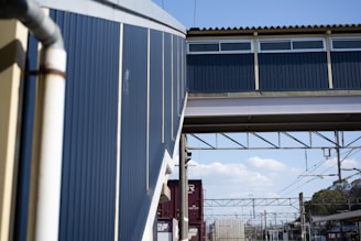 A railway station platform featuring an overhead pedestrian bridge made of metallic and wooden materials. The bridge is painted in blue and beige with windows at the top. A section of the bridge connects to the station building, which has metal siding. Below, train tracks run along the platform with power lines and shipping containers in the background. The sky is partly cloudy, creating a bright, industrial setting.