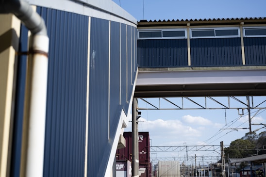 A railway station platform featuring an overhead pedestrian bridge made of metallic and wooden materials. The bridge is painted in blue and beige with windows at the top. A section of the bridge connects to the station building, which has metal siding. Below, train tracks run along the platform with power lines and shipping containers in the background. The sky is partly cloudy, creating a bright, industrial setting.