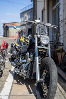 A close-up view of a row of motorcycles parked on a street, with the focus on the front of a black motorcycle featuring chrome detailing and wide handlebars. Behind it, other motorcycles with various colors and styles are visible. The scene is set against a backdrop of urban buildings under a clear blue sky.