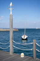A small sailboat is moored near a wooden dock, with a clear blue sky and the ocean in the background. A weather vane is prominently displayed in the foreground, and a red and a white lighthouse can be seen in the distance.