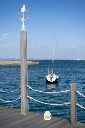 A small sailboat is moored near a wooden dock, with a clear blue sky and the ocean in the background. A weather vane is prominently displayed in the foreground, and a red and a white lighthouse can be seen in the distance.