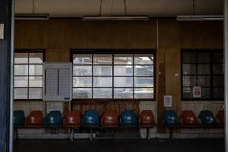 A dimly lit waiting area with a series of plastic chairs in alternating colors of teal and orange, facing a wall with large, multi-pane windows. A timetable or schedule is mounted on the wall. The interior has wooden paneling and visible fluorescent lights attached to the ceiling.