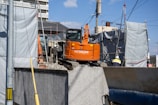 An orange Hitachi excavator is parked in a construction site surrounded by gray covered structures. A construction worker wearing a helmet stands near the vehicle. The background shows some residential or commercial buildings and a utility pole with overhead wires.