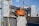 An orange Hitachi excavator is parked in a construction site surrounded by gray covered structures. A construction worker wearing a helmet stands near the vehicle. The background shows some residential or commercial buildings and a utility pole with overhead wires.