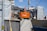 An orange Hitachi excavator is parked in a construction site surrounded by gray covered structures. A construction worker wearing a helmet stands near the vehicle. The background shows some residential or commercial buildings and a utility pole with overhead wires.