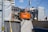 An orange Hitachi excavator is parked in a construction site surrounded by gray covered structures. A construction worker wearing a helmet stands near the vehicle. The background shows some residential or commercial buildings and a utility pole with overhead wires.