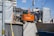 An orange Hitachi excavator is parked in a construction site surrounded by gray covered structures. A construction worker wearing a helmet stands near the vehicle. The background shows some residential or commercial buildings and a utility pole with overhead wires.
