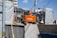 An orange Hitachi excavator is parked in a construction site surrounded by gray covered structures. A construction worker wearing a helmet stands near the vehicle. The background shows some residential or commercial buildings and a utility pole with overhead wires.