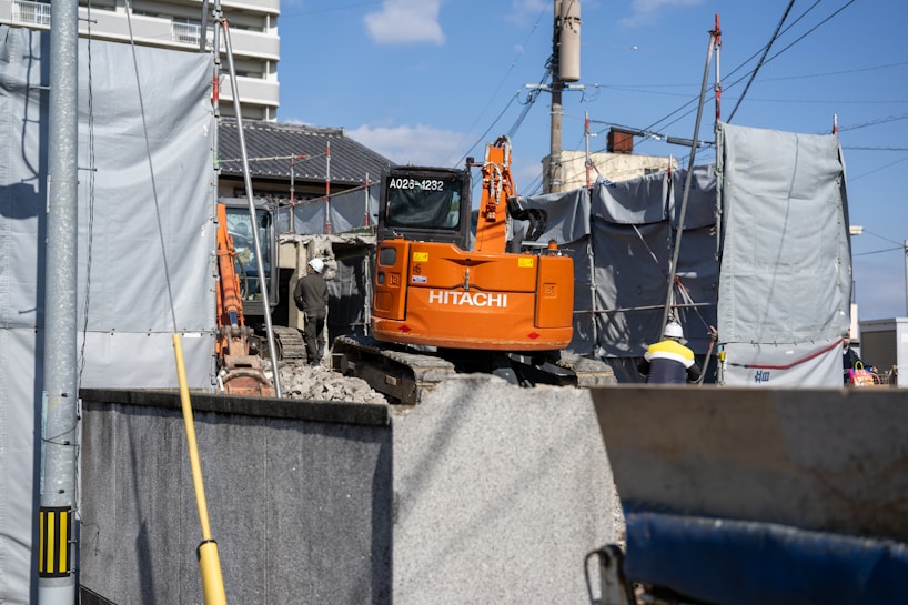 An orange Hitachi excavator is parked in a construction site surrounded by gray covered structures. A construction worker wearing a helmet stands near the vehicle. The background shows some residential or commercial buildings and a utility pole with overhead wires.
