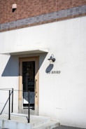 A minimalistic storefront with a simple door surrounded by a white wall. Above the door is a small overhang and an outdoor wall light. A tiled facade with shades of brown and gray tops the storefront. The name 'OARHINO' is displayed beside the door in black letters. A black metal railing accompanies the small set of steps leading to the entrance.
