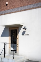 A minimalistic storefront with a simple door surrounded by a white wall. Above the door is a small overhang and an outdoor wall light. A tiled facade with shades of brown and gray tops the storefront. The name 'OARHINO' is displayed beside the door in black letters. A black metal railing accompanies the small set of steps leading to the entrance.