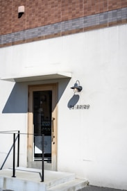 A minimalistic storefront with a simple door surrounded by a white wall. Above the door is a small overhang and an outdoor wall light. A tiled facade with shades of brown and gray tops the storefront. The name 'OARHINO' is displayed beside the door in black letters. A black metal railing accompanies the small set of steps leading to the entrance.