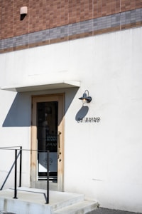 A minimalistic storefront with a simple door surrounded by a white wall. Above the door is a small overhang and an outdoor wall light. A tiled facade with shades of brown and gray tops the storefront. The name 'OARHINO' is displayed beside the door in black letters. A black metal railing accompanies the small set of steps leading to the entrance.
