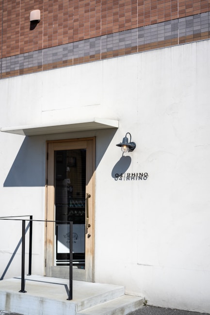 A minimalistic storefront with a simple door surrounded by a white wall. Above the door is a small overhang and an outdoor wall light. A tiled facade with shades of brown and gray tops the storefront. The name 'OARHINO' is displayed beside the door in black letters. A black metal railing accompanies the small set of steps leading to the entrance.