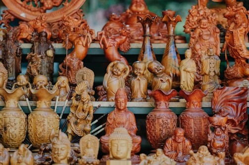 A collection of intricately carved wooden statues and vases, featuring various deities and traditional figures. The carvings display a high level of detail and craftsmanship, with smiling Buddhas, serene monks, and elaborate vases arranged closely together on a shelf.