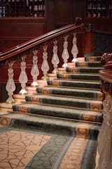 A grand staircase with ornate wooden banisters and intricately carved balusters. The steps are covered with a luxurious, patterned carpet in shades of green and beige. The rich wood tones of the railing contrast with the lighter colors of the marble steps and walls.