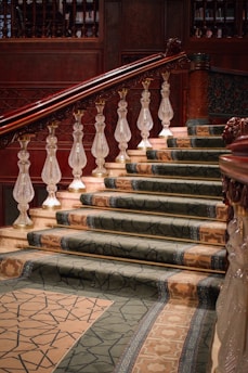 A grand staircase with ornate wooden banisters and intricately carved balusters. The steps are covered with a luxurious, patterned carpet in shades of green and beige. The rich wood tones of the railing contrast with the lighter colors of the marble steps and walls.