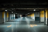 A dimly lit underground parking garage with yellow directional arrows on the floor and numbered parking spots along the sides. The walls are concrete with yellow and black markings, and fluorescent lights are evenly spaced across the ceiling.