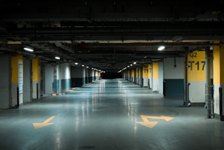 A dimly lit underground parking garage with yellow directional arrows on the floor and numbered parking spots along the sides. The walls are concrete with yellow and black markings, and fluorescent lights are evenly spaced across the ceiling.