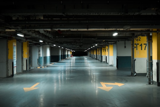 A dimly lit underground parking garage with yellow directional arrows on the floor and numbered parking spots along the sides. The walls are concrete with yellow and black markings, and fluorescent lights are evenly spaced across the ceiling.