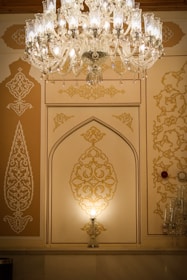 An ornate chandelier with intricate glasswork hangs from the ceiling, illuminating a wall with detailed golden patterns. The wall features artistic, geometric designs with a stylish arch in the center. The ambient light creates a warm and luxurious atmosphere.