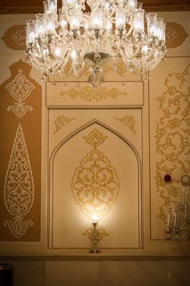 An ornate chandelier with intricate glasswork hangs from the ceiling, illuminating a wall with detailed golden patterns. The wall features artistic, geometric designs with a stylish arch in the center. The ambient light creates a warm and luxurious atmosphere.