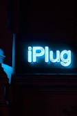 A neon sign brightly displays the word 'iPlug' against a dark background. The glowing blue lettering stands out prominently in the low light setting.