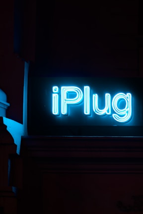 A neon sign brightly displays the word 'iPlug' against a dark background. The glowing blue lettering stands out prominently in the low light setting.