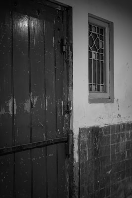 A black and white photograph depicting a rough wooden door with visible wear and an old window with metal bars. The wall next to the door and window shows signs of aging and decay, with peeling paint and cracked tiles.