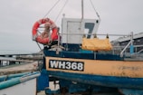 A small fishing boat is docked by the shore with its hull painted in blue and yellow, showing the registration number WH368. A bright orange lifebuoy with rope is attached to the side, and a yellow bag labeled as a safety ladder is secured to the railing. The background includes a pier extending into the water and some storage buildings.