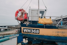 A small fishing boat is docked by the shore with its hull painted in blue and yellow, showing the registration number WH368. A bright orange lifebuoy with rope is attached to the side, and a yellow bag labeled as a safety ladder is secured to the railing. The background includes a pier extending into the water and some storage buildings.