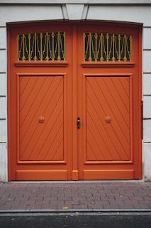 A pair of large, double wooden doors painted in a vibrant orange color. The doors feature a diagonal plank pattern and are adorned with decorative black and gold ironwork at the top. The surrounding wall is made of a smooth, light-colored stone.
