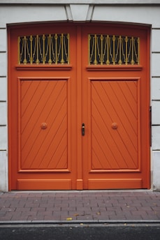 A pair of large, double wooden doors painted in a vibrant orange color. The doors feature a diagonal plank pattern and are adorned with decorative black and gold ironwork at the top. The surrounding wall is made of a smooth, light-colored stone.