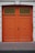 A pair of large, double wooden doors painted in a vibrant orange color. The doors feature a diagonal plank pattern and are adorned with decorative black and gold ironwork at the top. The surrounding wall is made of a smooth, light-colored stone.