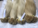 Bundles of long, straight, blonde hair extensions neatly arranged and secured with yellow bands. Each bundle has a label detailing its specifications, and they are placed on a soft white fur surface.