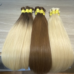 Three bundles of straight hair lay side by side on a wooden surface. Each bundle is neatly tied with yellow bands. The colors of the hair range from light blonde to brown.