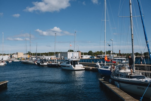 A marina with several boats and yachts docked along the wooden piers, surrounded by calm water under a clear blue sky. Some boats are equipped with tall masts and flags, while people are seen standing on one of the boats. Behind the marina, various industrial buildings and green trees are visible.