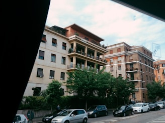 Several mid-rise apartment buildings line a street, with trees and parked cars in the foreground. The architecture features balconies and various window styles, suggesting a residential area. The sky is partly cloudy, casting a soft light over the scene.