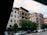 Several mid-rise apartment buildings line a street, with trees and parked cars in the foreground. The architecture features balconies and various window styles, suggesting a residential area. The sky is partly cloudy, casting a soft light over the scene.