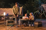 A nighttime camping scene features three people enjoying an outdoor meal. Two people are seated at a table, engaging in conversation while holding drinks. A third person stands beside a portable grill, cooking food. String lights create a cozy ambiance, and various camping equipment is visible, including a portable power station, cooler, and folding tables.
