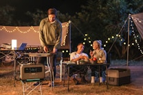A nighttime camping scene features three people enjoying an outdoor meal. Two people are seated at a table, engaging in conversation while holding drinks. A third person stands beside a portable grill, cooking food. String lights create a cozy ambiance, and various camping equipment is visible, including a portable power station, cooler, and folding tables.