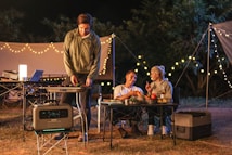 A nighttime camping scene features three people enjoying an outdoor meal. Two people are seated at a table, engaging in conversation while holding drinks. A third person stands beside a portable grill, cooking food. String lights create a cozy ambiance, and various camping equipment is visible, including a portable power station, cooler, and folding tables.