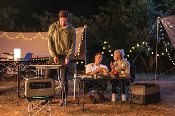 A nighttime camping scene features three people enjoying an outdoor meal. Two people are seated at a table, engaging in conversation while holding drinks. A third person stands beside a portable grill, cooking food. String lights create a cozy ambiance, and various camping equipment is visible, including a portable power station, cooler, and folding tables.