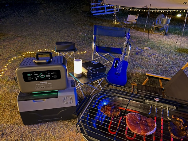A nighttime outdoor scene featuring a portable power station and cooler on the ground next to a folding chair. A grill with meat is situated in the foreground. A blue acoustic guitar leans against a chair under a canopy with string lights, creating a cozy atmosphere.
